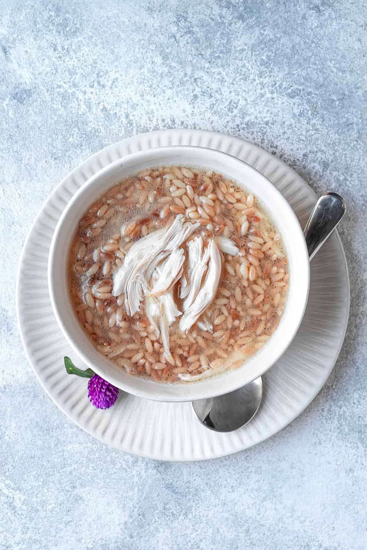 overhead image of Middle Eastern Orzo Soup topped with shredded chicken with a white bowl on top of a white plate with a spoon on the side
