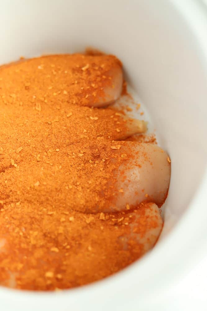 close up image of 4 raw chicken breasts in a white crock pot covered with fajita seasoning
