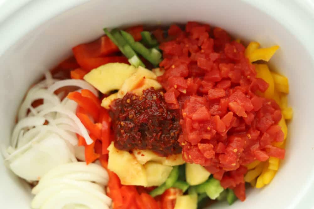 close up image of a white crock pot full of chicken, bell peppers, white onions, pineapple, tomatoes, and chipotle peppers to make chicken fajitas