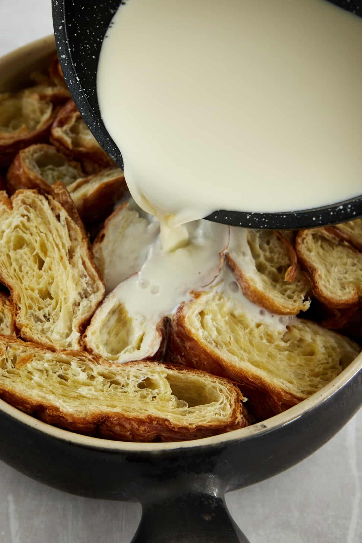 a sweetened milk and heavy cream mixture being poured over pieces of croissants in a round dish