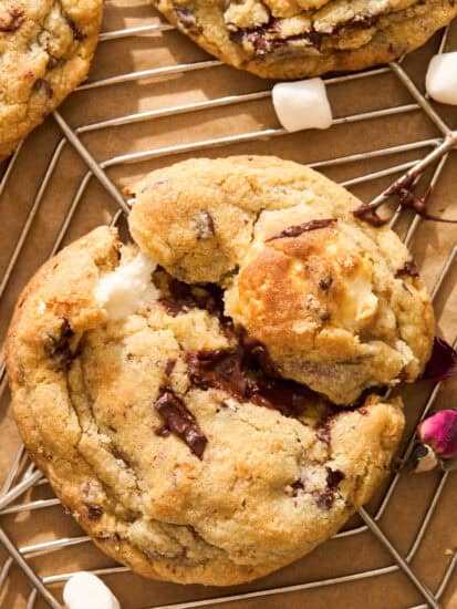 A close-up of chocolate chip cookies with melted chocolate and marshmallows on a cooling rack, with mini marshmallows scattered nearby.