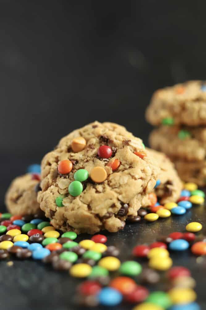 a close up image of a monster cookie leaning on two other monster cookies with a stack in the background and m&ms scattered around