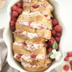 challah french toast cut hasselback and stuffed with raspberry cheesecake filling in a white baking dish with raspberries on the side