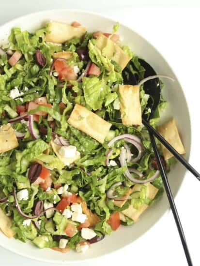overhead image of Greek Salad in a large white bowl with serving utensils on the side