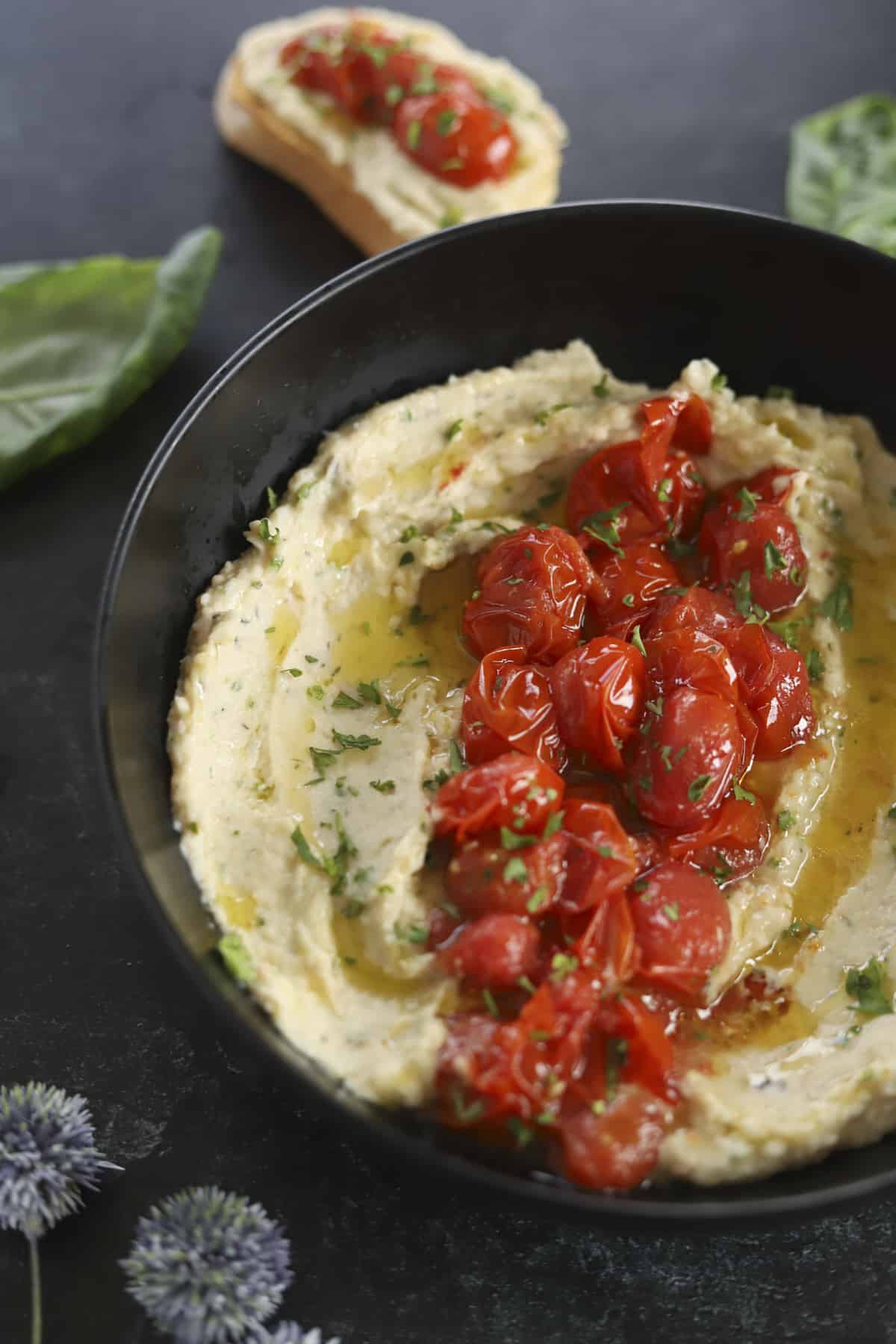 bowl of white bean dip with roasted tomatoes and baguette on side.