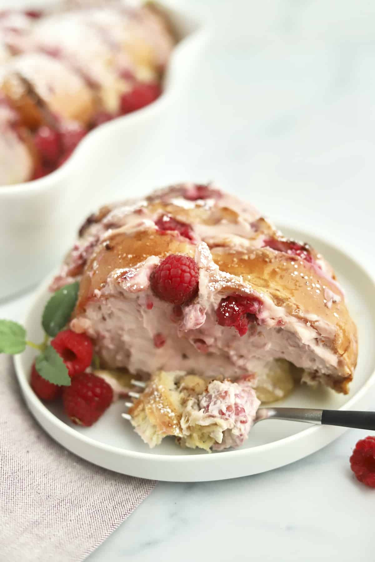 a serving of raspberry cheesecake challah french toast on a plate with fresh raspberries and a fork taking a bite