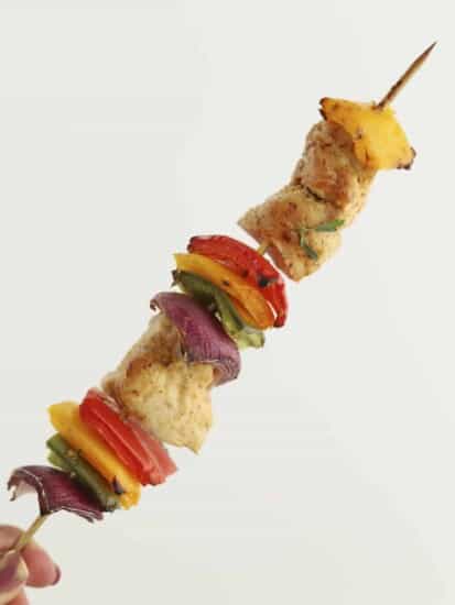 a hand holding an oven-baked chipotle chicken skewer with red, yellow, and green peppers and red onion