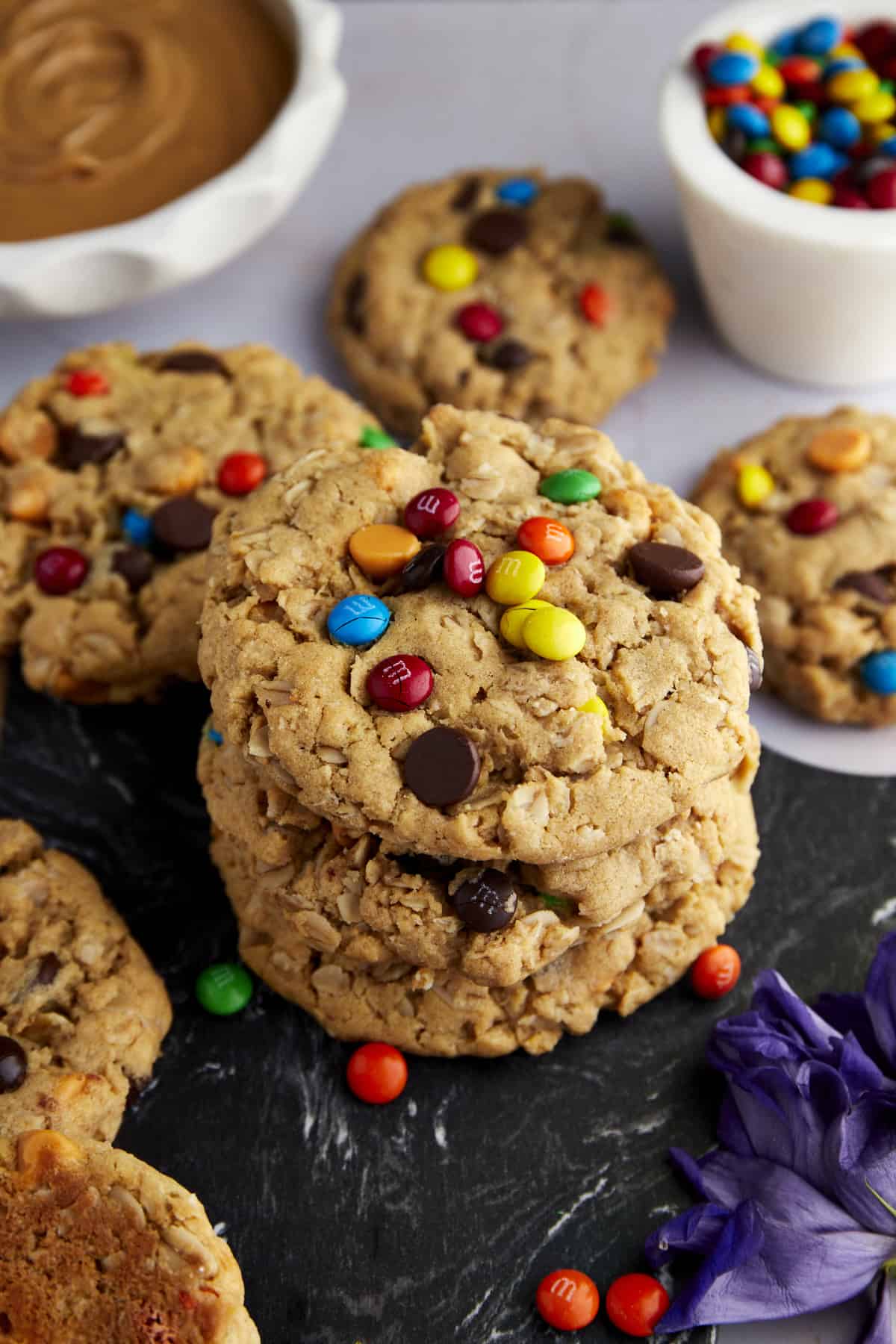 Monster cookies topped with M&M's and chocolate chips with bowls of peanut butter and M&M's in the background
