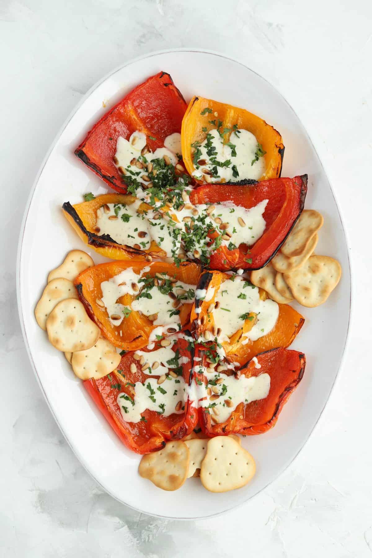 overhead image of a white serving platter full of quartered roasted peppers and crackers that have been drizzled with a whipped feta recipe and topped with pine nuts and chopped parsley