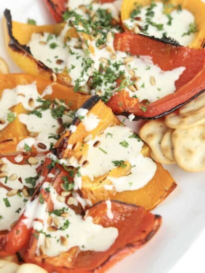 a white serving dish full of quartered roasted peppers and crackers topped with whipped feta, pine nuts, and chopped parsley