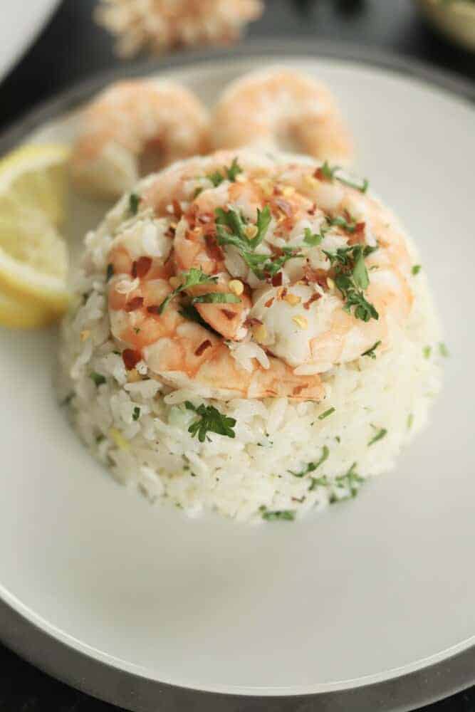 close up image of lemon garlic shrimp and rice on a white plate topped with fresh herbs with two shrimp and a lemon wedge in the background