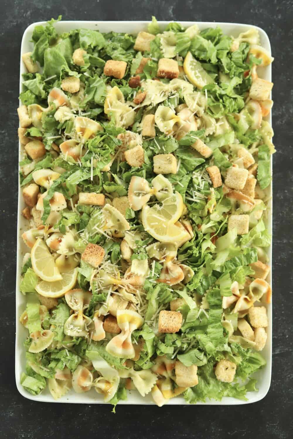 a rectangular white serving platter full with pesto pasta salad made with lettuce, noodles, Parmesan, croutons, and a pesto sauce topped with lemon wedges
