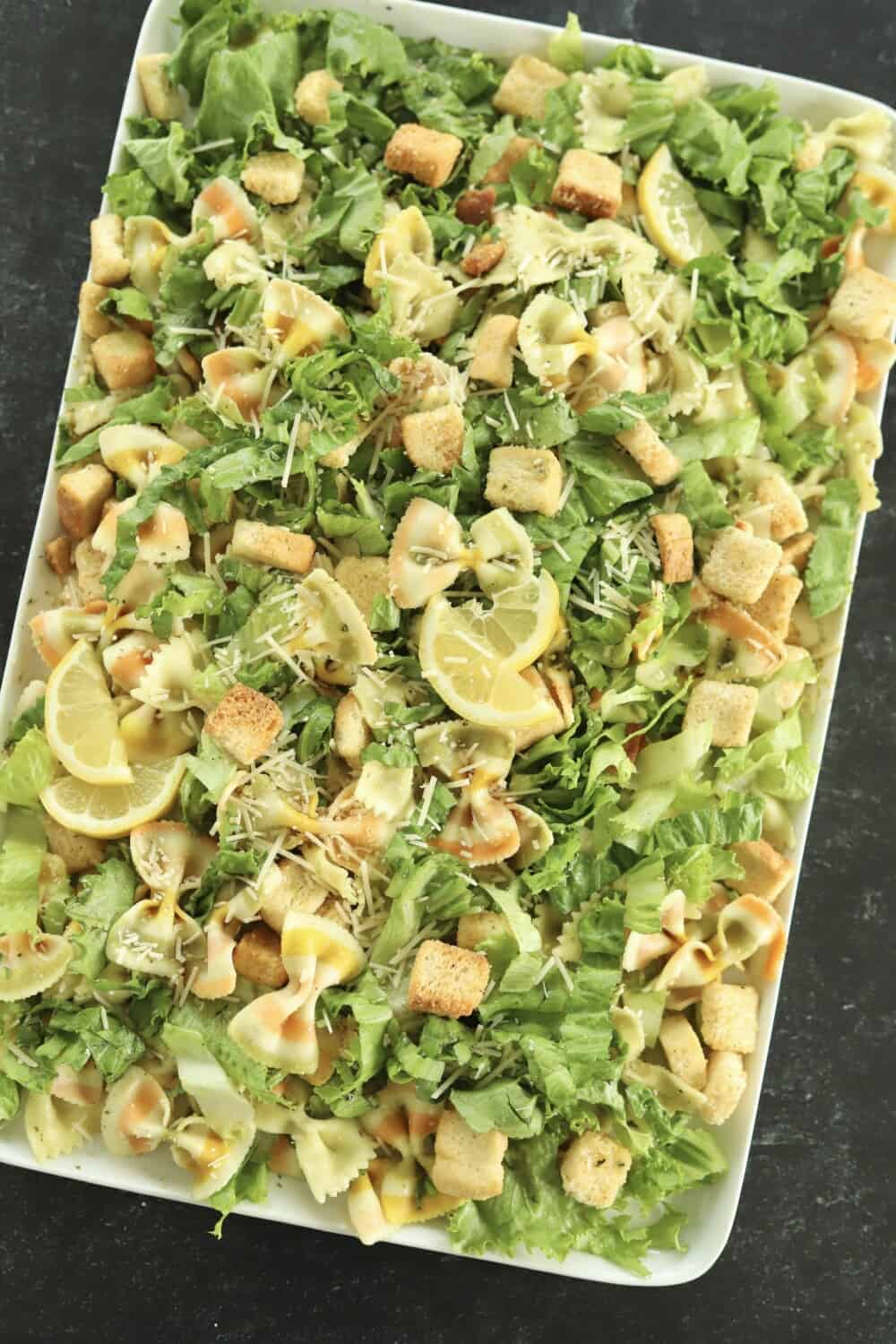 overhead image of a white serving platter full of pesto pasta salad made with lettuce, noodles, croutons, and Parmesan