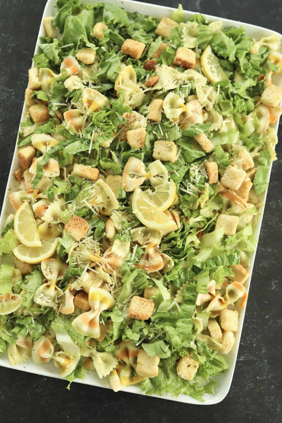 overhead image of a white serving platter full of pesto pasta salad made with lettuce, noodles, croutons, and Parmesan