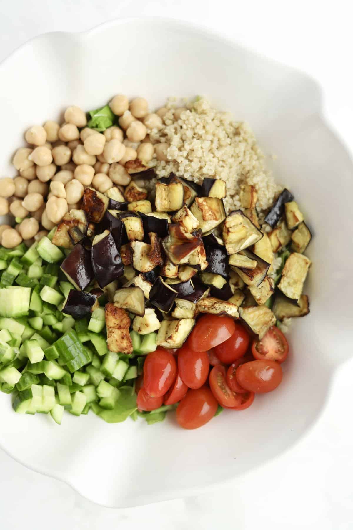 overhead image of quinoa, chickpeas, roasted eggplant, cucumber, and tomatoes in a bowl ready to be combined for roasted eggplant salad
