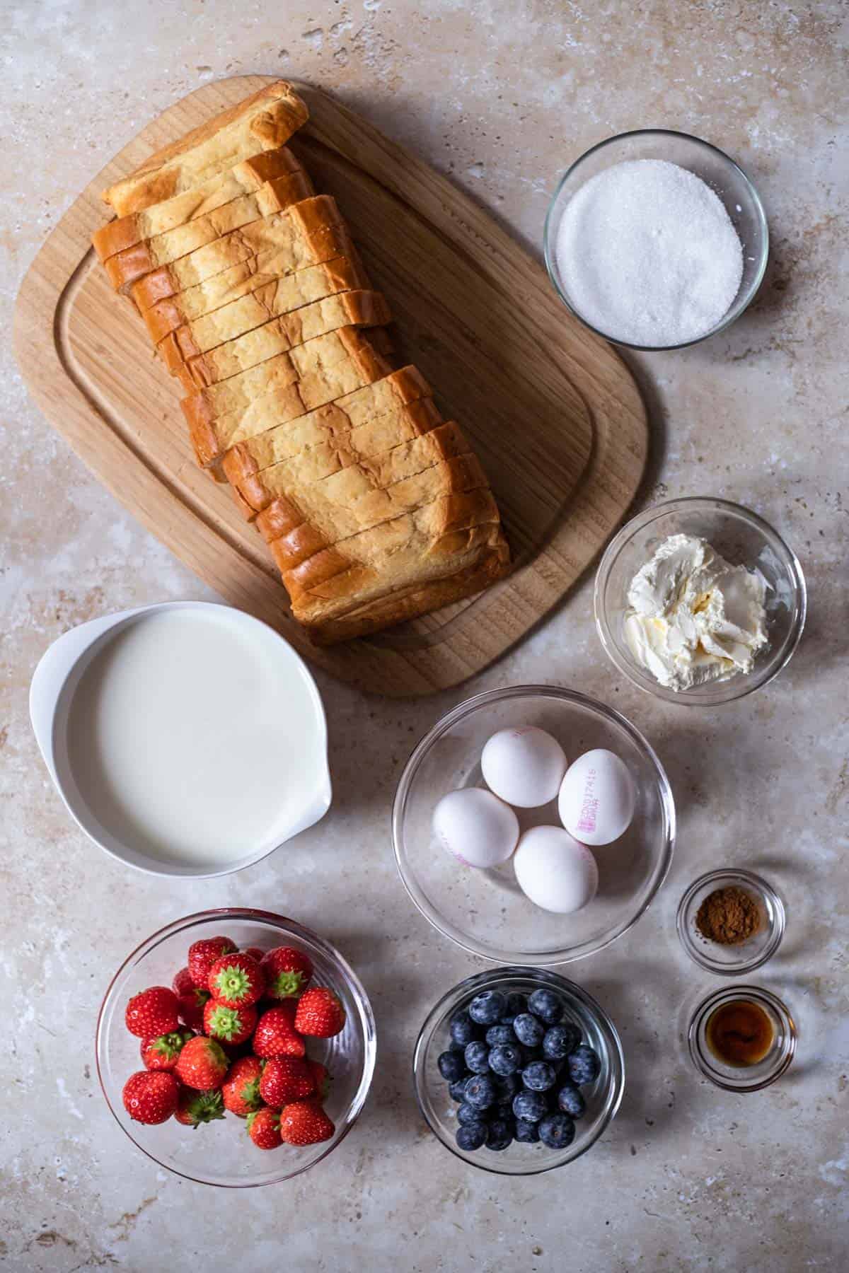 a loaf of sliced bread, sugar, cream cheese, cinnamon, eggs, milk, strawberries, and blueberries