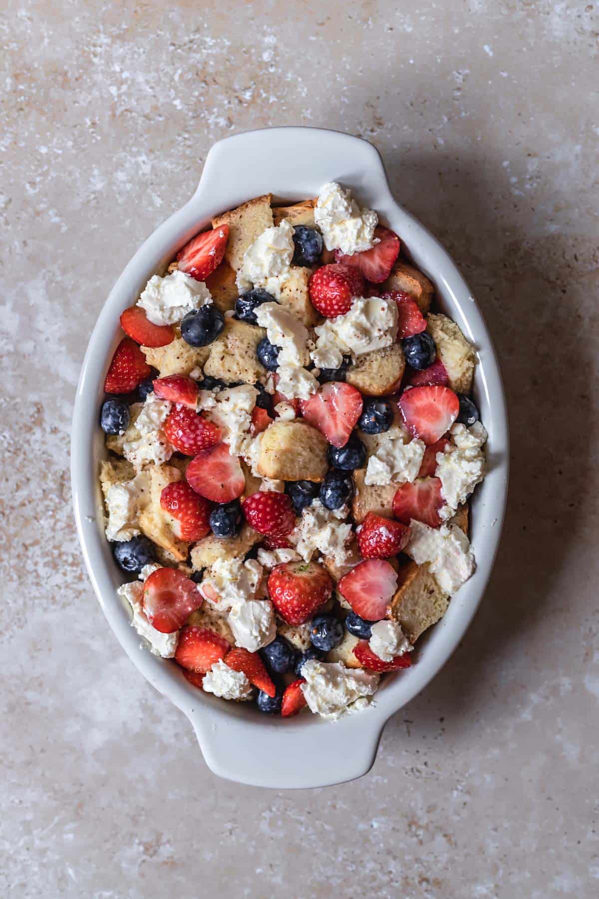 bread pieces, sliced strawberries, blueberries, and cream cheese in an oval baking dish