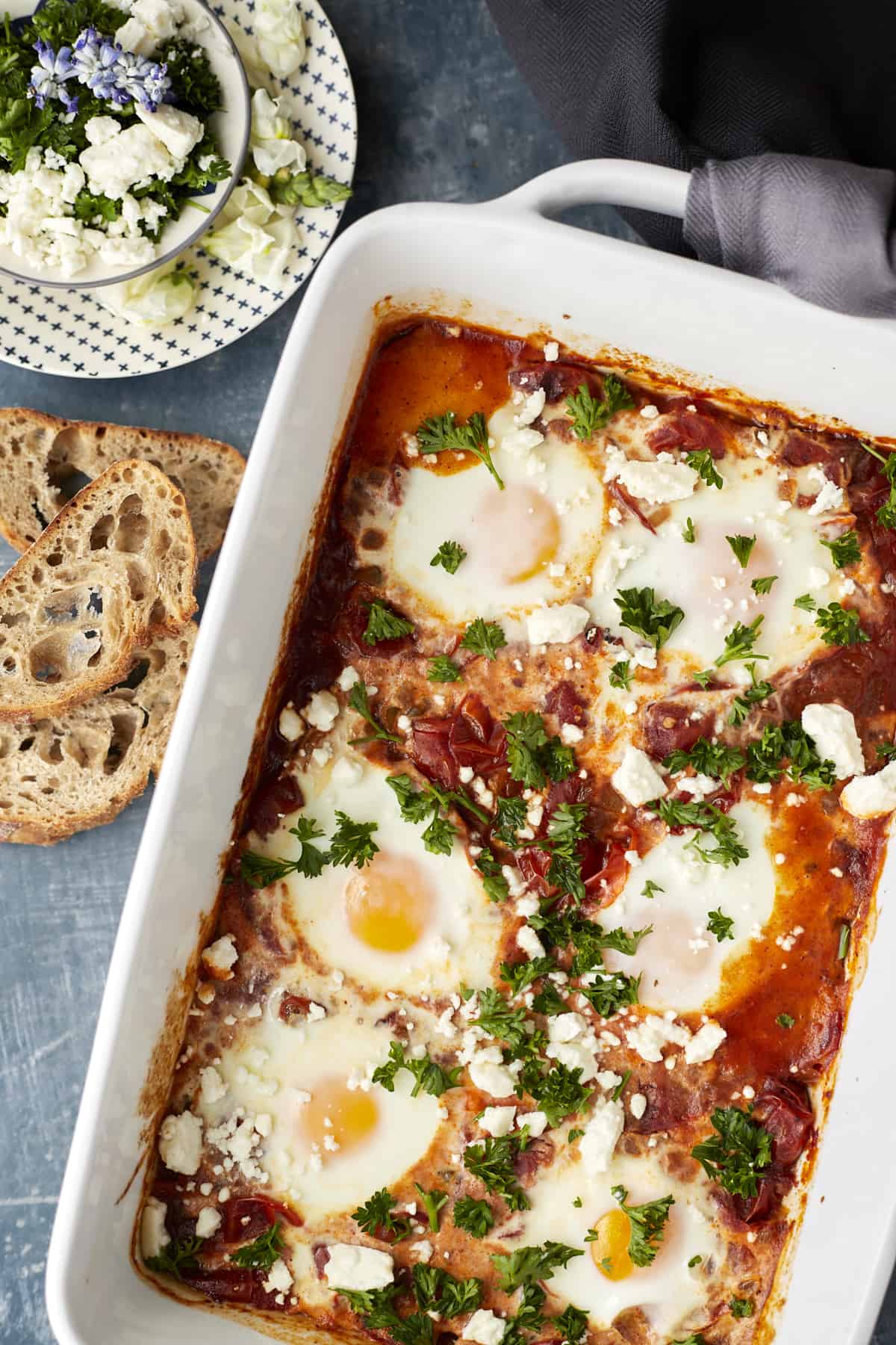 closeup overhead of oven baked shakshuka in a baking dish garnished with feta cheese and parsley. Shown with a side of toasted crostini and a plate with extra feta cheese and parsley