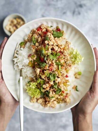 two hands holding a plate full of asian chicken with white rice.