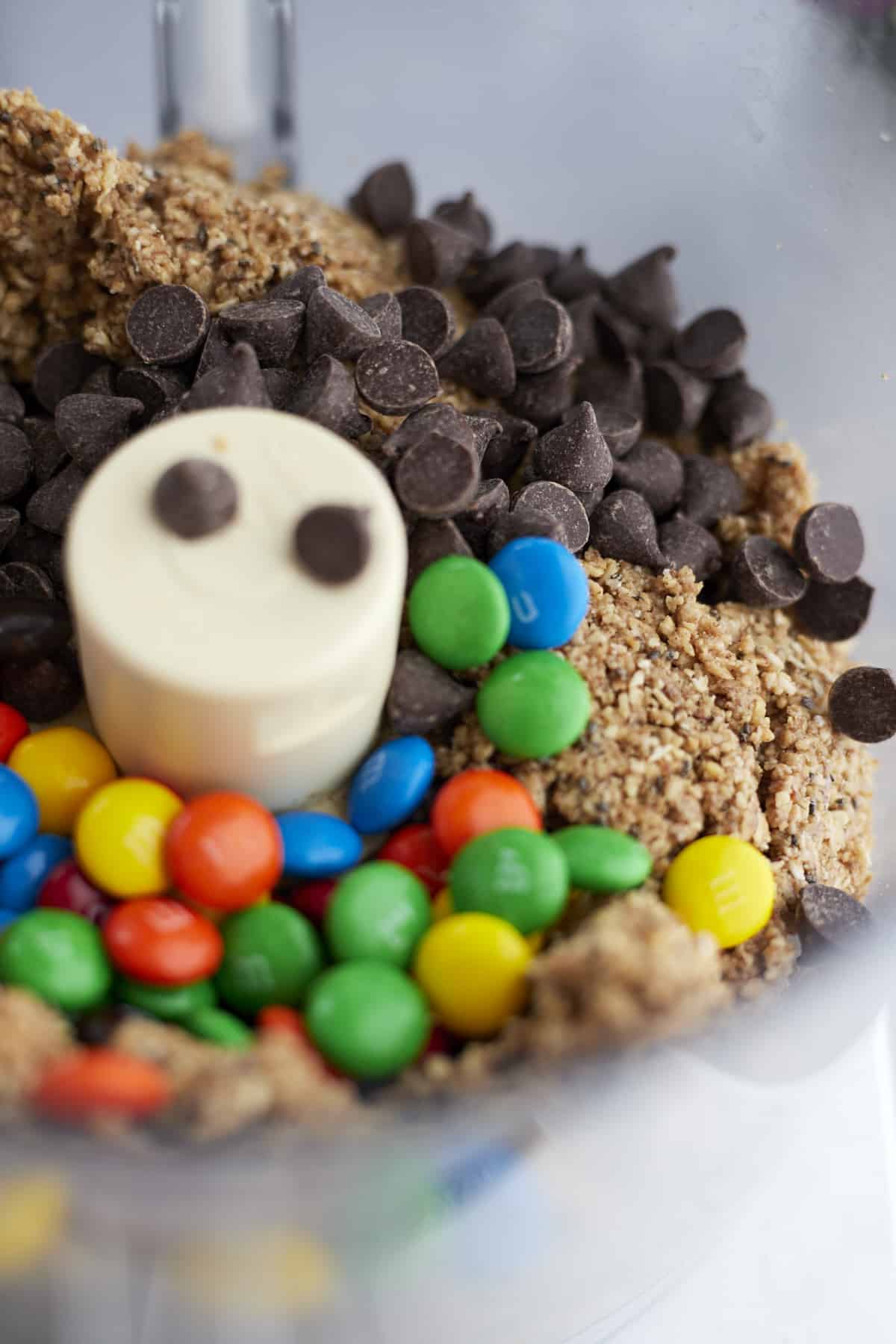 close up image of a mixture of ground oats and almonds, M&Ms, and chocolate chips in a food processor