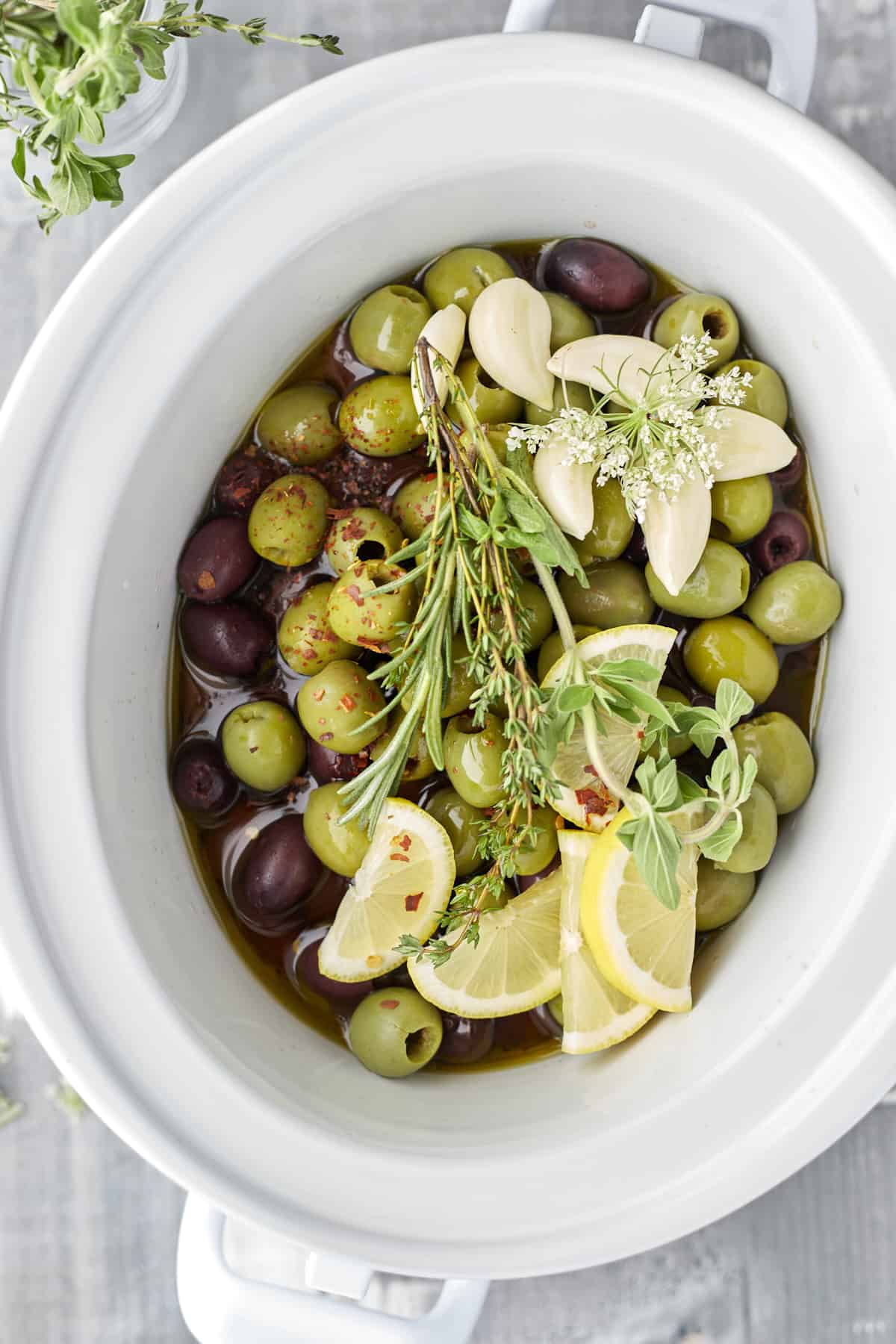 overhead image of green and red olives, lemon wedges, garlic, and herbs in a slow cooker to make roasted olives