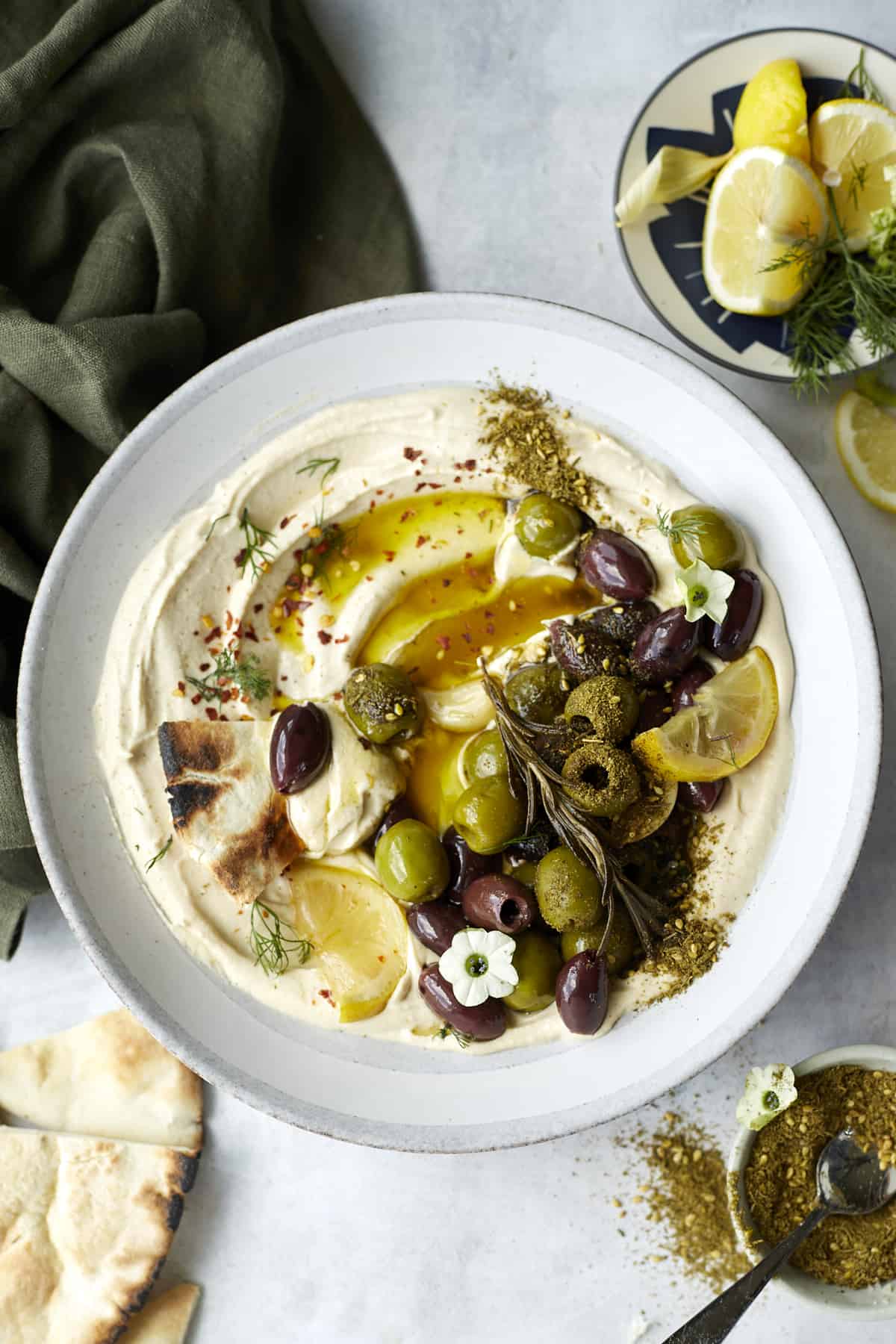overhead image of a white plate with hummus topped with roasted olives, herbs, and seasonings