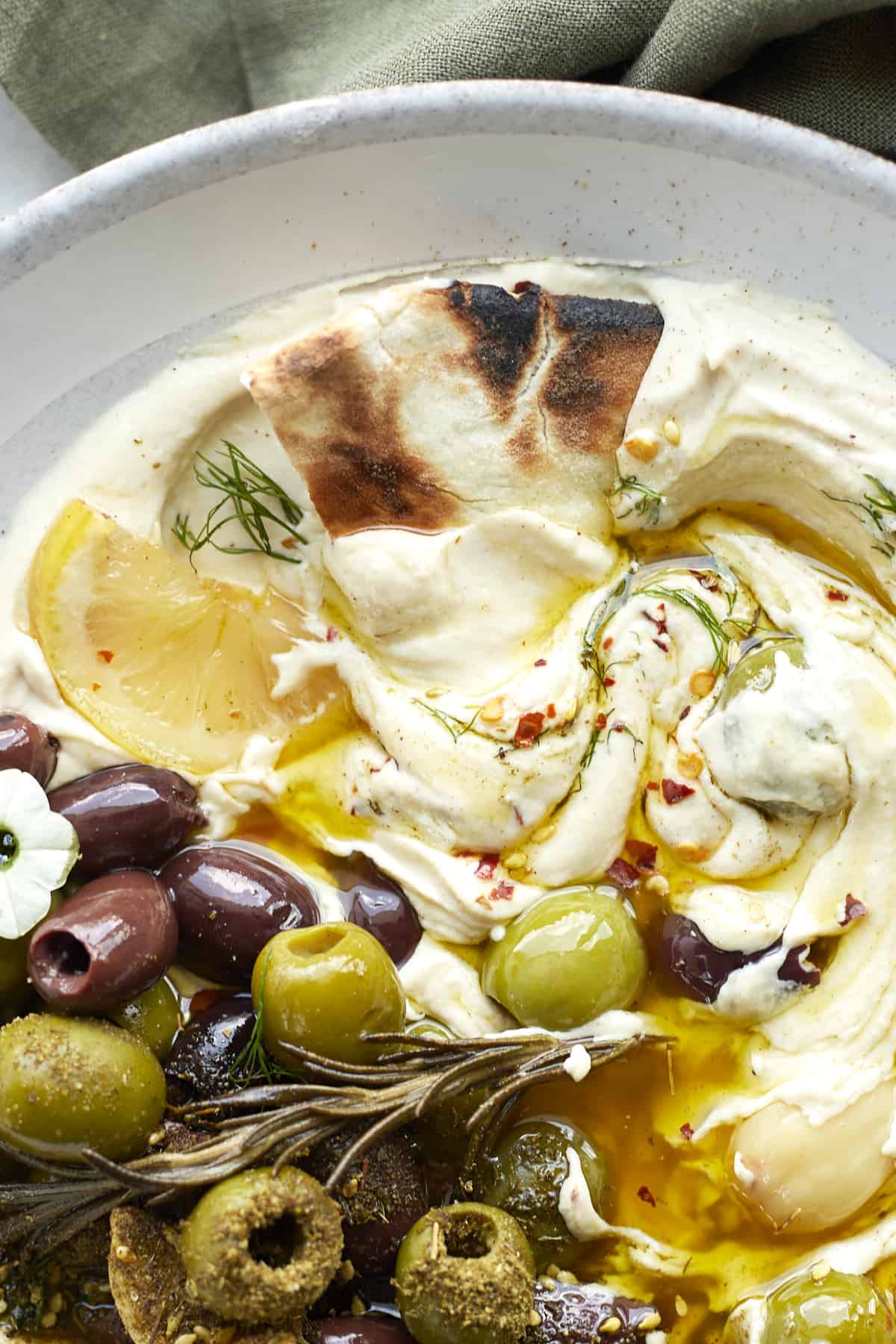 close up image of hummus topped with roasted olives, herbs, and lemons, and pita bread dipping in