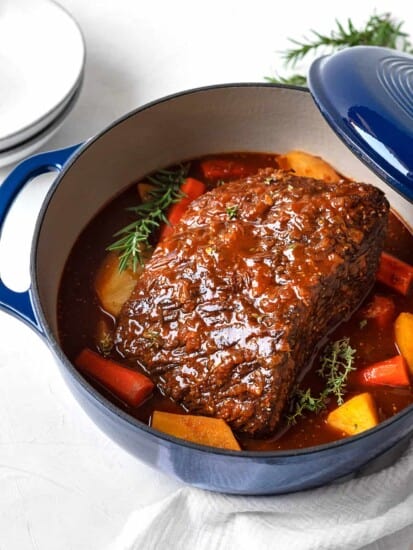 a fully cooked beef pot roast, onions, carrots, potatoes, and gravy in a blue dutch oven