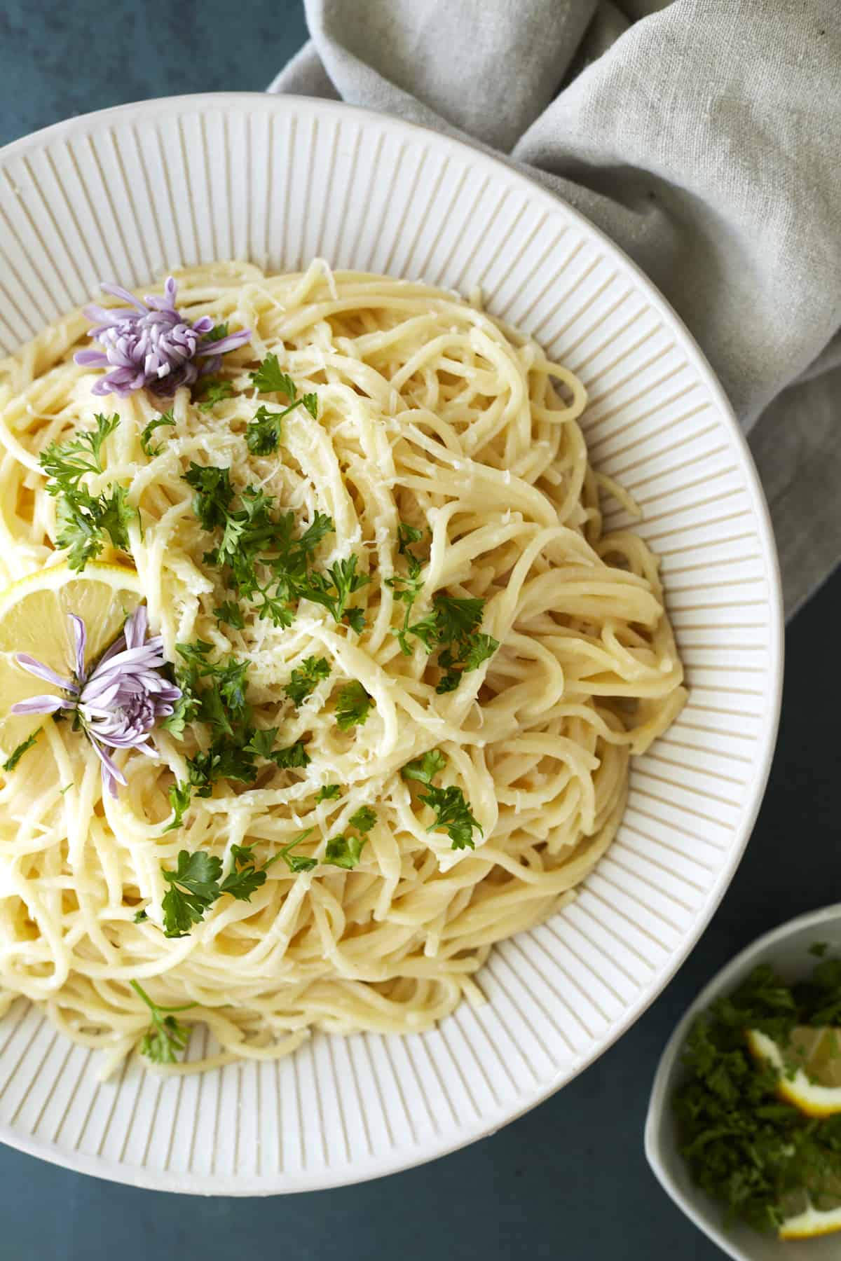 overhead image of a white plate full of lemon pasta topped with fresh herbs, purple flowers, and a lemon wedge