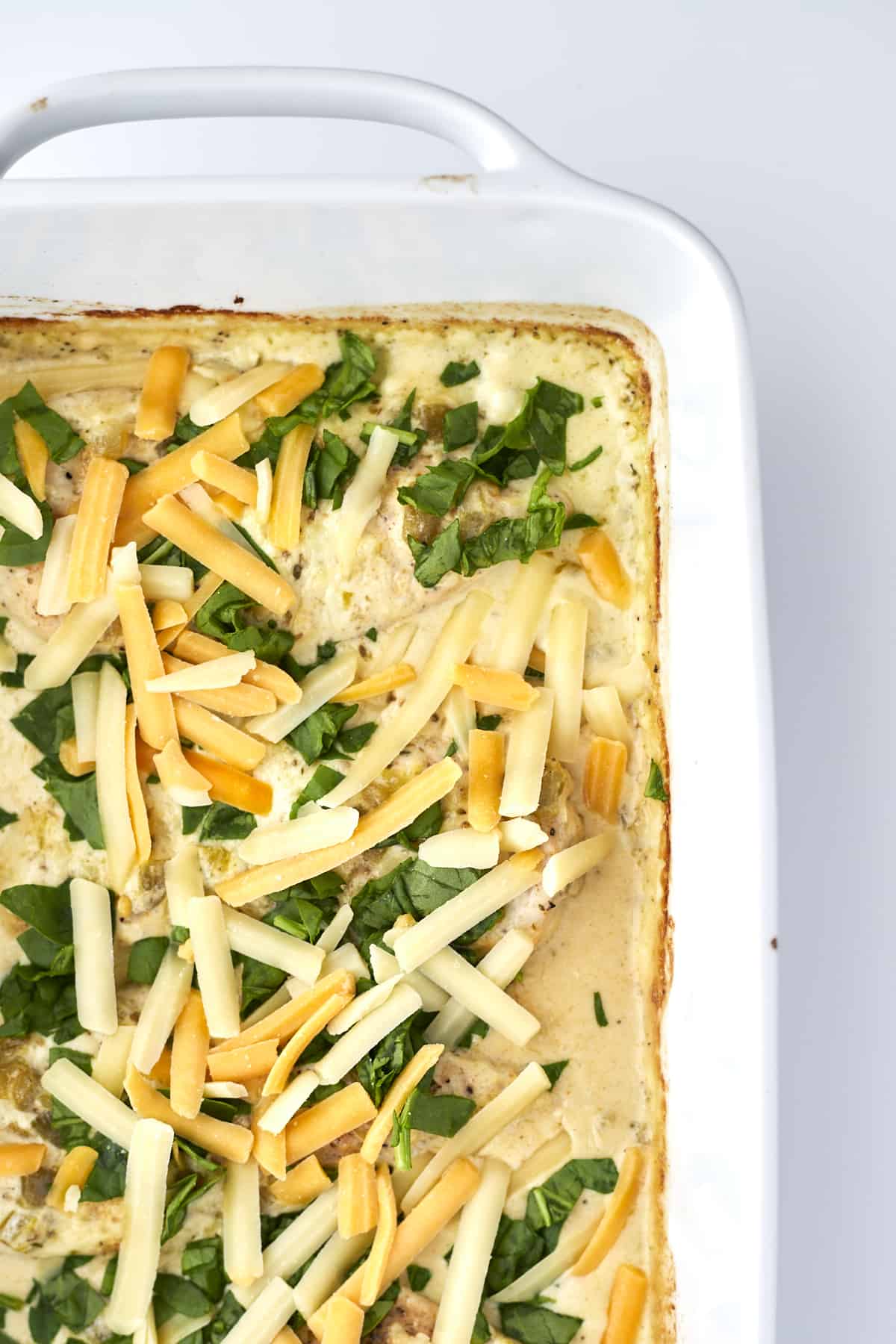 overhead image of unbaked cheesy chicken in a white casserole dish topped with spinach and shredded cheese