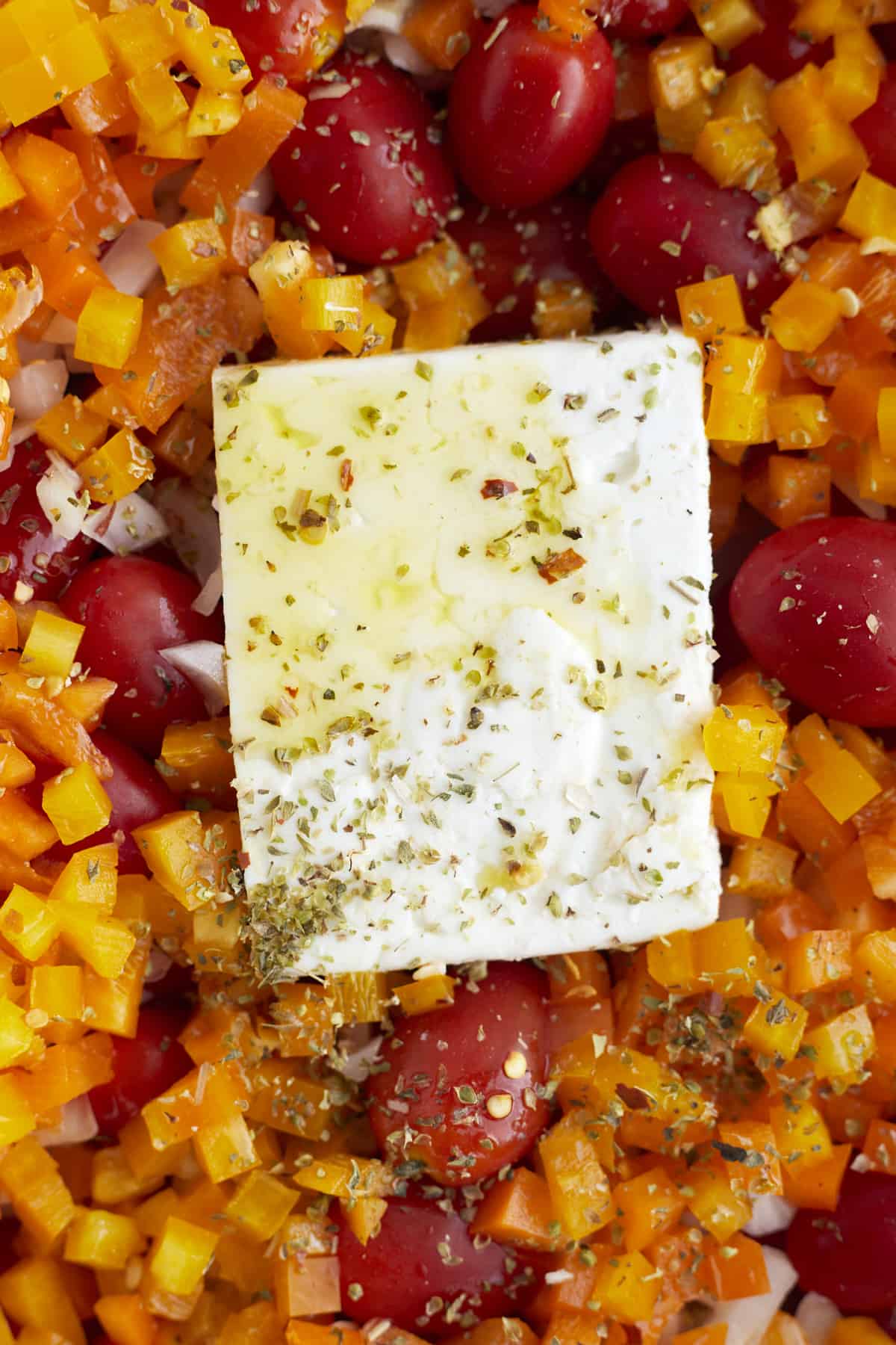 a close up image of chopped peppers, onions, tomatoes, and a block of feta cheese topped with olive oil and seasonings.