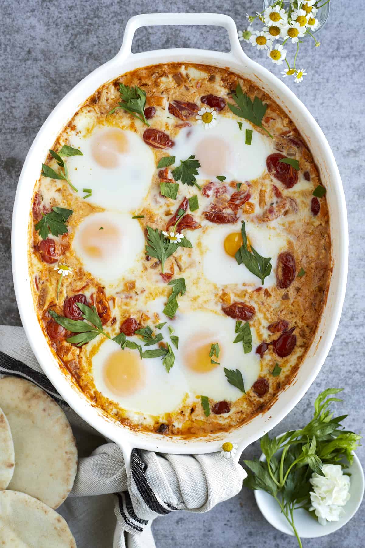 overhead image of 6 oven baked eggs in a white baking dish surrounding by vegetables and melted feta cheese topped with fresh herbs.