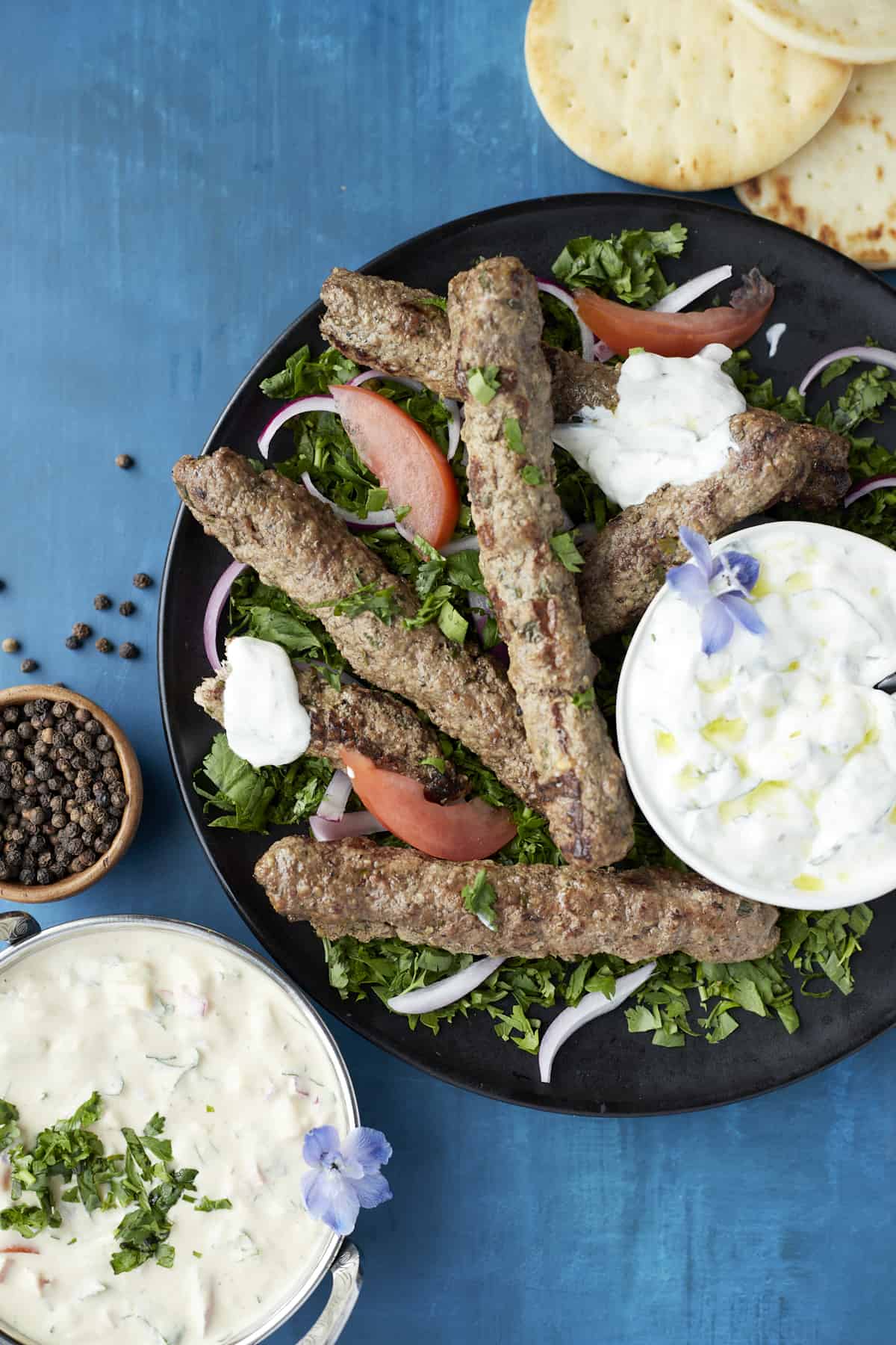 overhead image of beef kofta on top of a salad with bowls of tzatziki, a small bowl of capers, and pita on the side