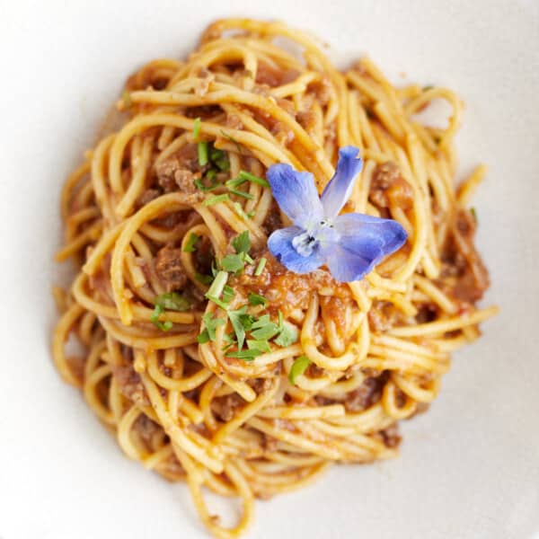 overhead image of a white bowl with a serving of one pot spaghetti with ground beef topped with fresh herbs and a blue flower.