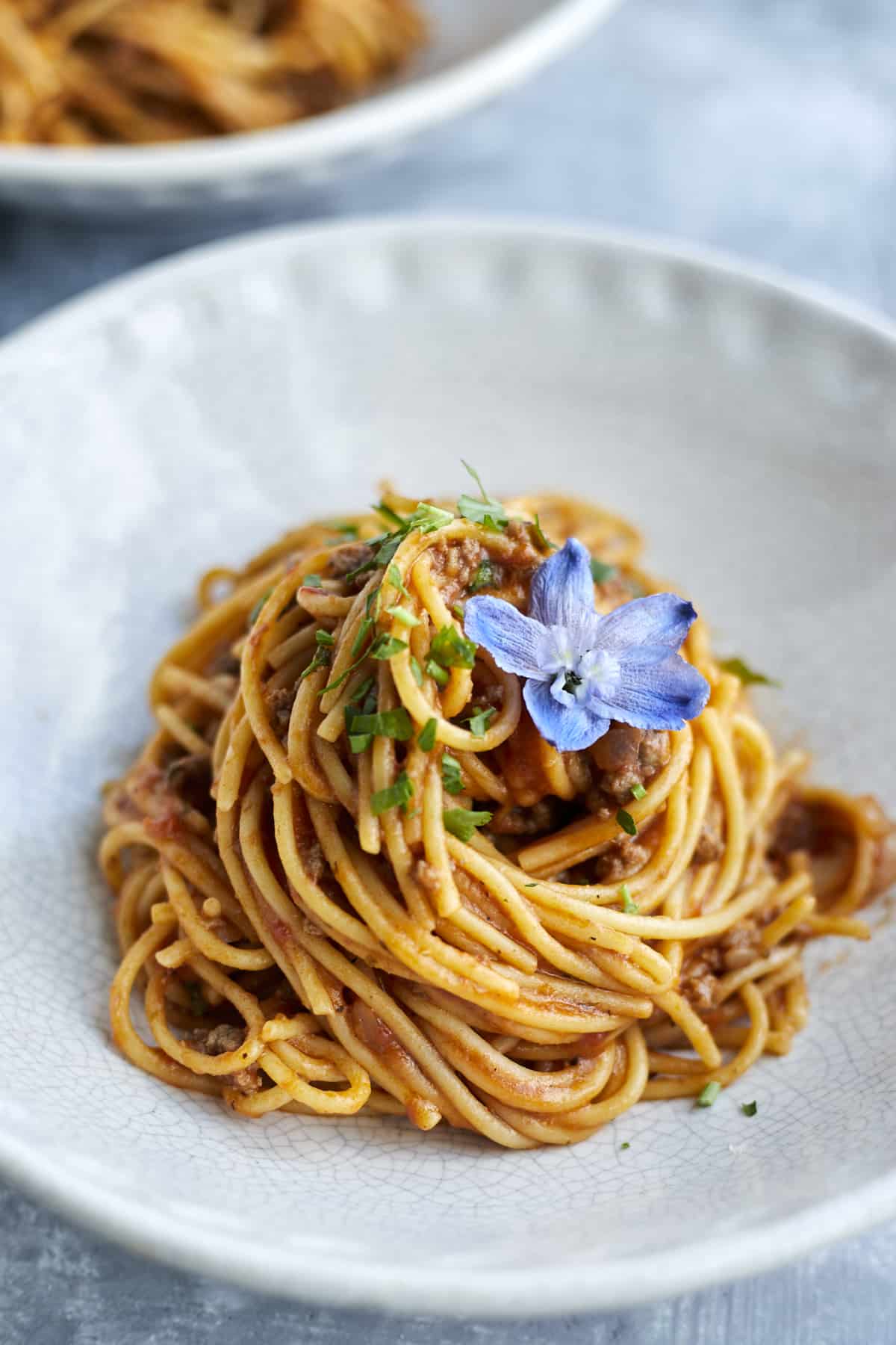 a close up image of a white plate with a serving on one pot spaghetti with ground beef topped with fresh herbs and a blue flower.