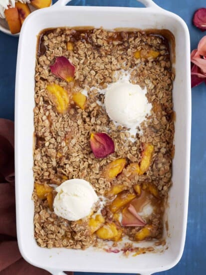 overhead image of a baked peach crisp topped with two scoops of vanilla ice cream with a serving missing out of the bottom righthand corner
