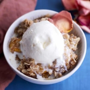a small white bowl full of peach crisp topped with a scoop of vanilla ice cream