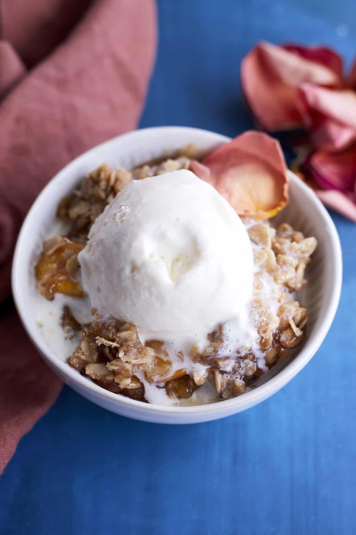 a small white bowl full of a serving of peach crisp topped with slightly melted vanilla ice cream.