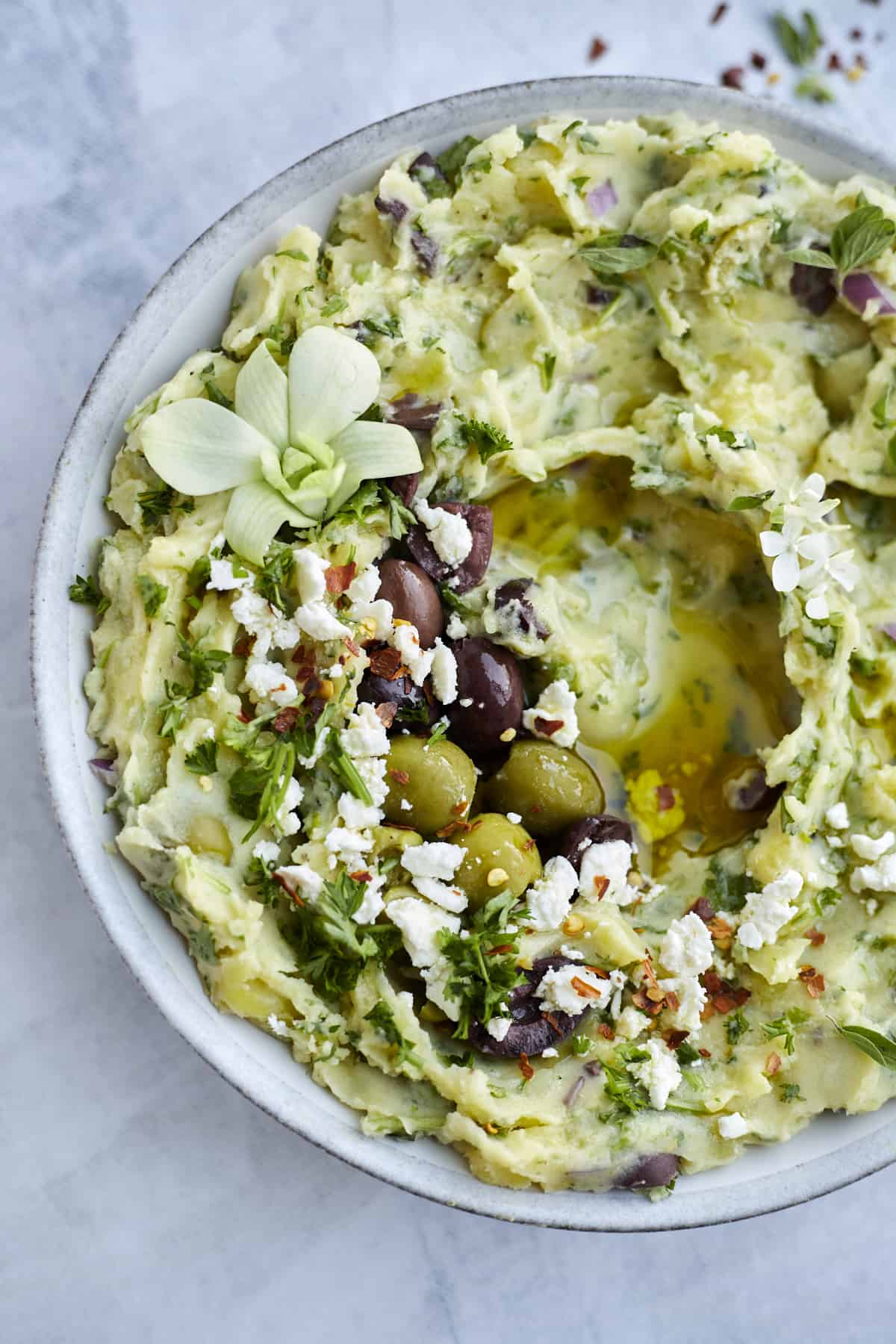 overhead image of a white bowl full of Greek-Inspired Yukon Gold Mashed Potatoes topped with olive oil, kalamata olives, green olives, feta cheese, red pepper flakes, and fresh herbs.