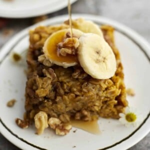 a piece of baked pumpkin oatmeal on a white plate topped with walnuts and banana coins with a drizzle of maple syrup being poured on top