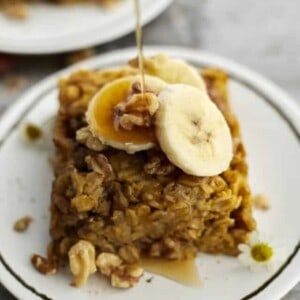 a piece of baked pumpkin oatmeal on a white plate topped with walnuts and banana coins with a drizzle of maple syrup being poured on top