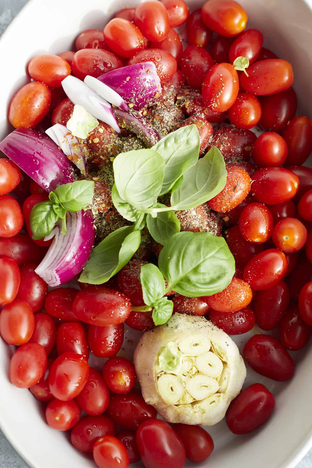 a baking dish with cherry tomatoes, a whole head of garlic, red onion, shallot, basil, and seasonings