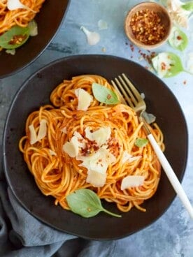 A serving of arrabiata sauce spaghetti on a black plate topped with fresh Parmesan cheese, basil leaves, and red pepper flakes with a fork on the side.