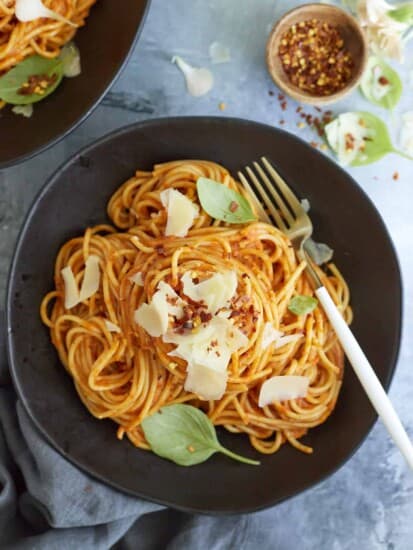 A serving of arrabiata sauce spaghetti on a black plate topped with fresh Parmesan cheese, basil leaves, and red pepper flakes with a fork on the side.