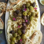 overhead image of a butter board topped with green and black olives, pine nuts, and seasonings with crostini around the sides
