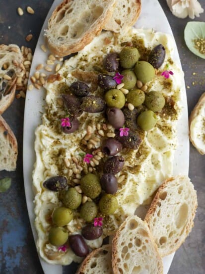 overhead image of a butter board topped with green and black olives, pine nuts, and seasonings with crostini around the sides