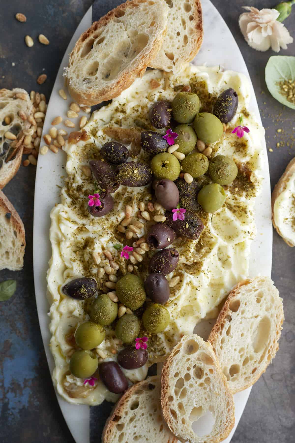 a white board topped with butter, green and black olives, and pine nuts with crostini around the sides