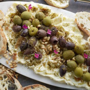a white board topped with butter, olives, pine nuts, and spices with bread and serving utensils along the edges