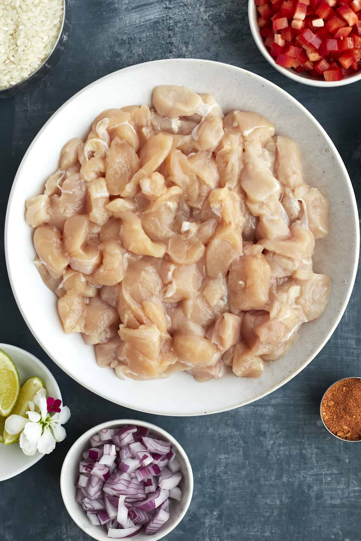 overhead image of a bowl of raw cubed chicken, chopped red onions, chopped red pepper, lime wedges, and uncooked white rice