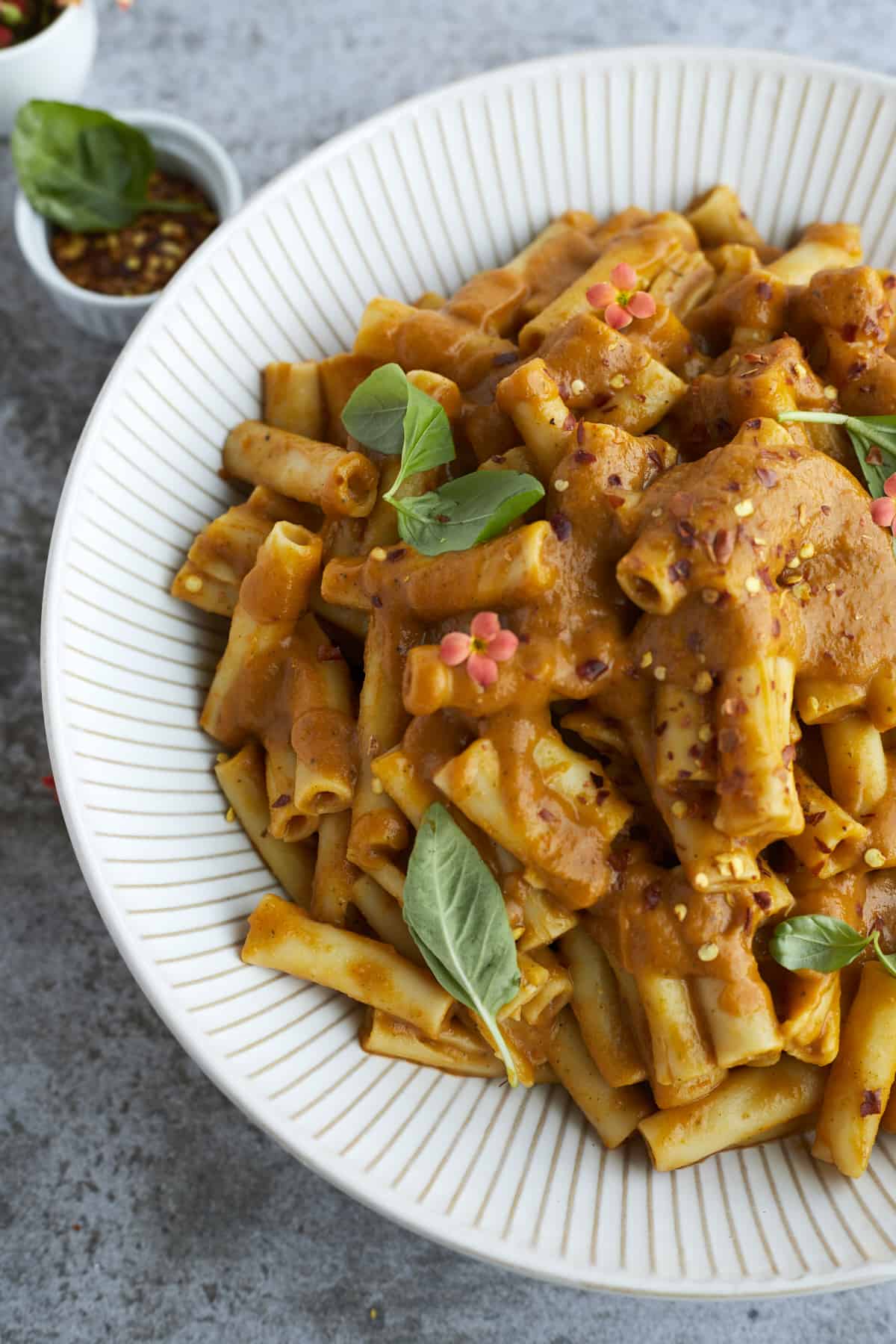 close up image of a white bowl full of cooked veggie pasta topped with fresh herbs and red pepper flakes